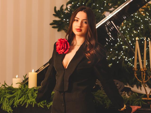 ChristinaBliss with long brown hair, wearing a black blazer with a red rose, exposing her tits, standing in front of a decorated Christmas tree, candles, and greenery.