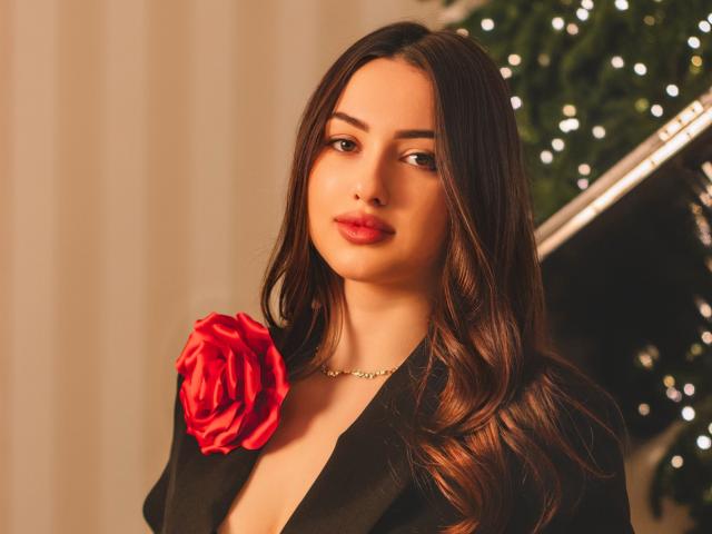ChristinaBliss with long brown hair, red rose on black jacket, red lips, subtle makeup, gold necklace, serious expression, Christmas tree with lights in background.