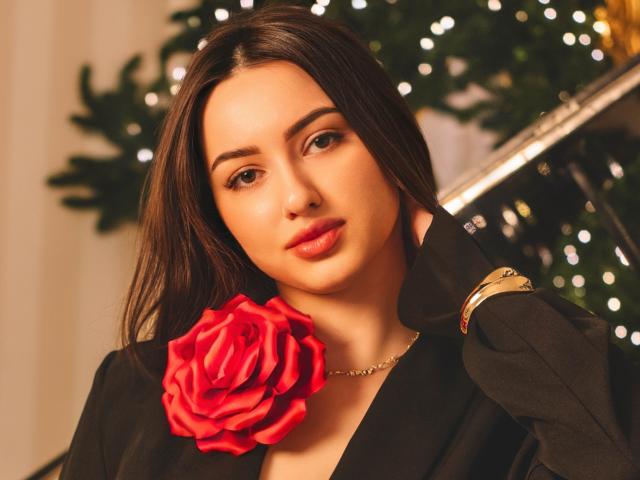 ChristinaBliss with long brown hair, wearing a black shirt, has a red rose pinned to her chest and a gold bracelet on her wrist, looking seductively at the camera.