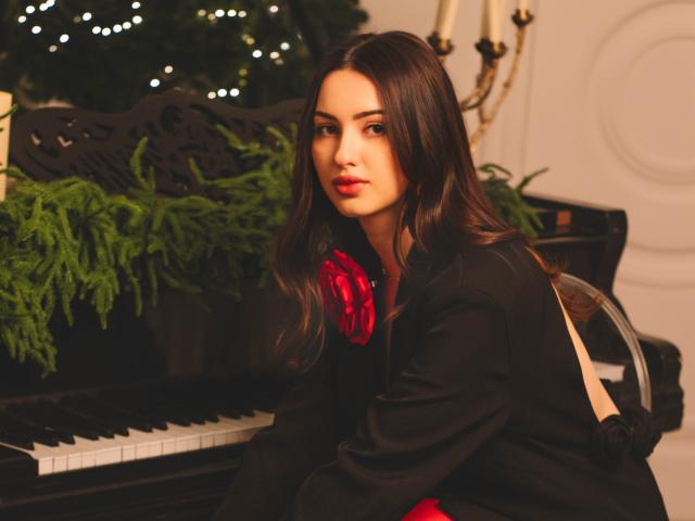ChristinaBliss sits in front of a black piano adorned with green ferns and Christmas lights. She has long, dark brown hair and red lipstick. She wears a black coat with a red bow on the left side. Her expression is neutral. The piano's detailed black carvings are visible. The image uses warm lighting. ChristinaBliss's face is slightly turned towards the camera. The overall composition is balanced and elegant.