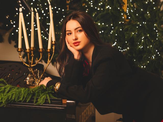 ChristinaBliss met lang bruin haar, een zwarte top, met haar hoofd rustend op haar hand, kijkend naar de camera, naast een gouden kandelaar met witte kaarsen, groene varens, een kerstboom met witte lichtjes..