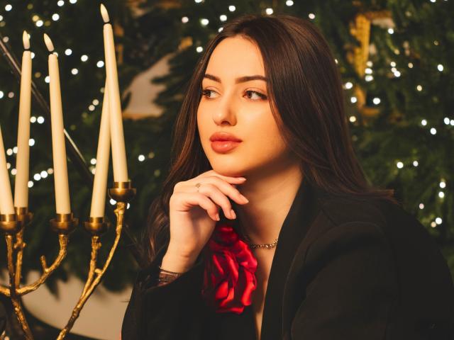 ChristinaBliss, com cabelo castanho escuro, segurando uma rosa vermelha na mão, usando um blazer preto, com um candelabro dourado, iluminação suave, olhando para o lado com os lábios vermelhos levemente entreabertos..