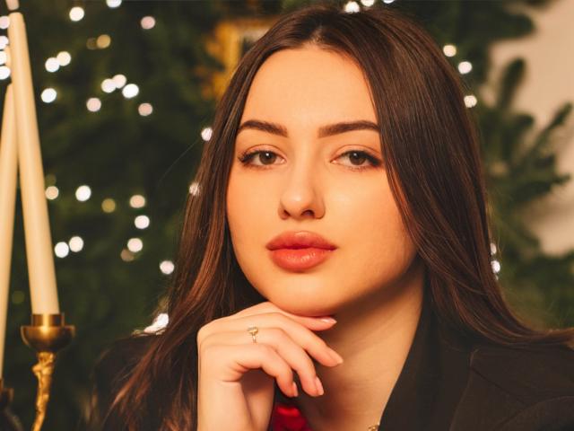 ChristinaBliss, met lang bruin haar, volle lippen en een neusring, kijkt naar de camera, met haar hand rustend op haar kin, gekleed in een zwarte jas, met kaarsen op de achtergrond..