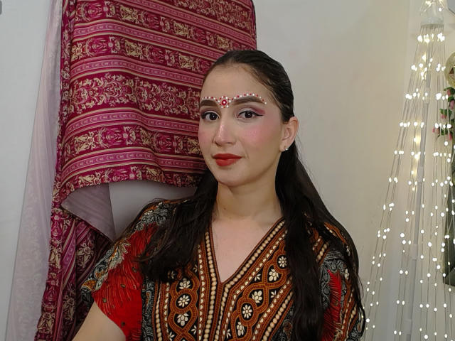 JalimaMona with red lipstick, dark hair in pigtails, wearing a red and gold patterned dress, and a jeweled forehead piece, standing in front of a pink and red curtain.