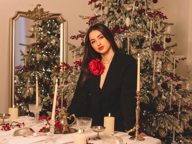 ChristinaBliss with long dark hair, red rose on black dress, standing by decorated Christmas tree, candles, and silverware on white table, looking cute with red lips and subtle makeup.