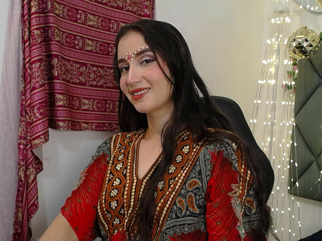 JalimaMona with long dark hair, red and black patterned dress, pearl headpiece, smiling, showing small tits, sitting in chair, red and pink patterned curtain, string lights in background.