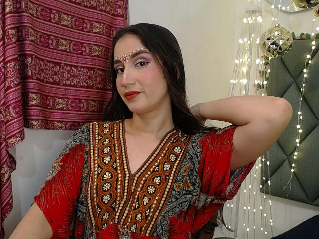JalimaMona with dark hair, red and brown patterned dress, white bead headpiece, arm raised, red lipstick, and patterned curtains. She's got a cute ass and a pretty face.