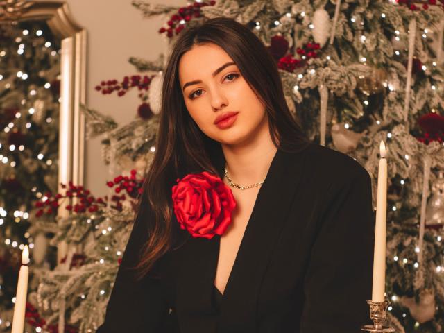 ChristinaBliss with long brown hair, wearing a black dress with a red rose on the chest, looking at the camera, standing in front of decorated Christmas trees with candles.