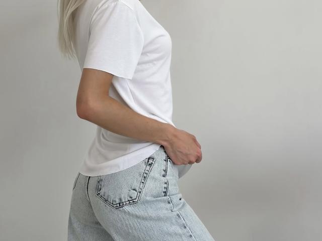GlariyaHot wearing a white tee and light blue denim jeans, side profile, showing a slim waist and hand in pocket, blonde hair, white background.