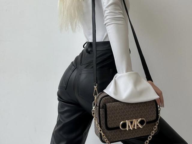 GlariyaHot's ass in black leather pants, white long-sleeve top, holding a brown Chanel purse with gold chain and "CM" logo, long blonde hair, white wall background.