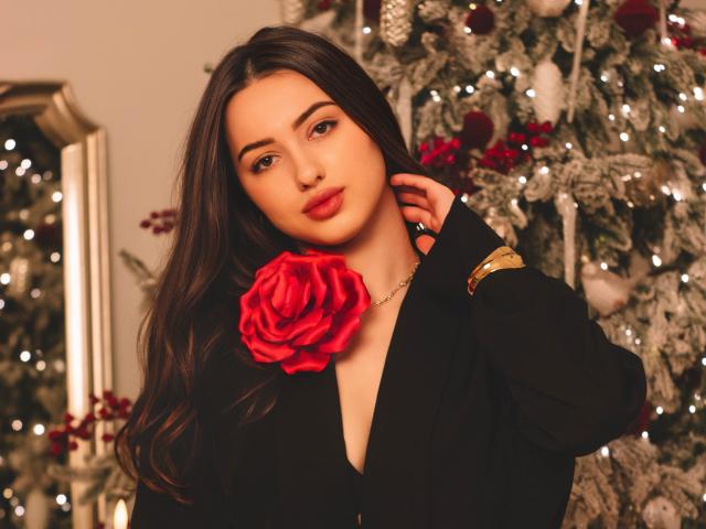 ChristinaBliss, with long dark brown hair, wears a black V-neck top, a gold bracelet on her left wrist, and a large red rose pinned to her chest. Her brown eyes and red lips stand out. She gazes directly at the camera with a slight, confident smile. The background features a decorated Christmas tree with white snow-like ornaments and red berries. The image has a warm, soft light, highlighting ChristinaBliss's smooth, fair skin. Her relaxed, yet elegant pose is central to the composition.