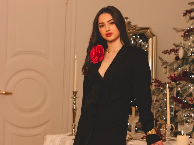 ChristinaBliss with long dark hair, wearing a black suit, red rose on chest, gold necklace, standing, looking at camera, warm lighting, Christmas tree with lights in background.