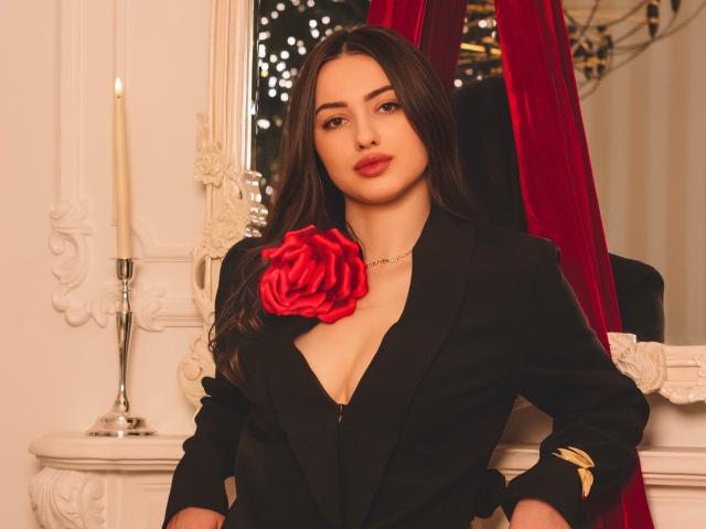 ChristinaBliss with long dark hair, wearing a black blazer, deep V-neck, red rose on chest, hands on hips, gold bracelet, red velvet curtain, white ornate mirror, candle.