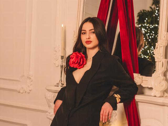 ChristinaBliss with long dark hair, wearing a black blazer, red rose on chest, red curtains behind, candle on white ornate mirror, looking at camera, hands in pockets.