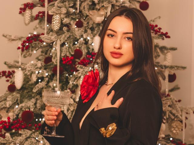 ChristinaBliss with long brown hair, red lipstick, black dress, gold button, holding a glass, standing in front of a Christmas tree with red ornaments and white snow.