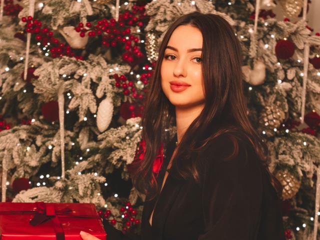 ChristinaBliss with long brown hair, red lipstick, and black top, holding a red gift box, standing in front of a Christmas tree with white lights and red berries.