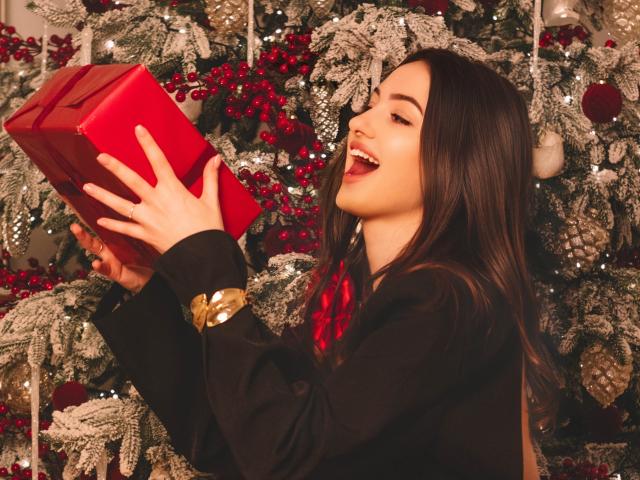 ChristinaBliss with long brown hair, black sweater, and gold bracelet, smiling while holding a red gift box, her tongue playfully sticking out. Christmas tree with red berries in background.