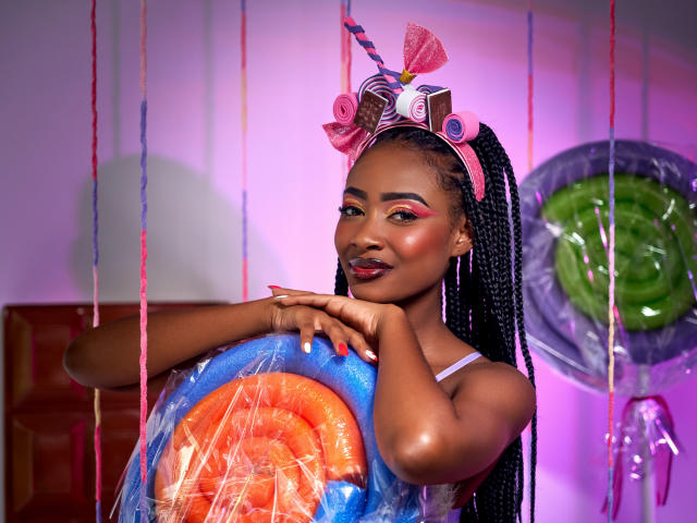 DalilaQueen, an African woman with dark skin and long braided hair, stands holding a large, colorful lollipop. She wears a pink headband with curly and spiral decorations. Her makeup includes pink eyeshadow and red lipstick. She has red nail polish. The lollipop is blue with an orange swirl, partially covered by clear plastic. A green lollipop is in the background. Bright, pink-purple lighting. Slick, shiny skin. Her expression is calm and confident. The image has a vibrant, playful feel.