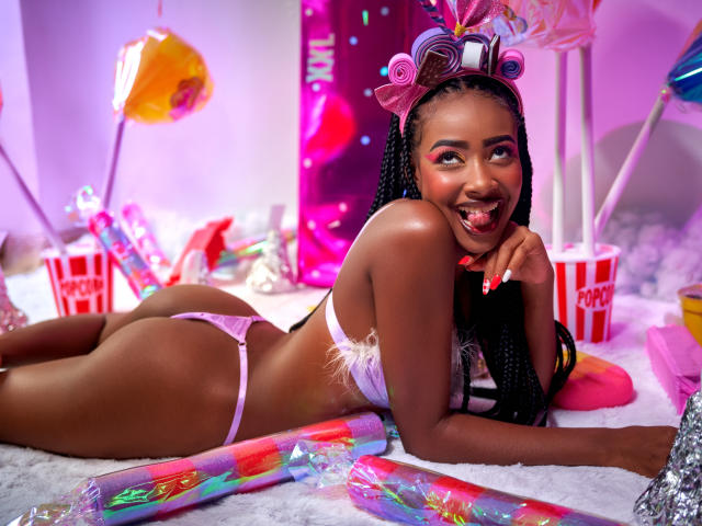 DalilaQueen, a dark-skinned woman with braids, lies on a white furry surface. She wears a pink lace thong and matching bra, with her tongue playfully sticking out. Her right hand rests on her chin. She has pink and purple eye makeup, and a colorful hair roller set on her head. The background is a mix of pink and purple, with colorful balloons and a red "popcorn" bucket. The image has bright, playful colors and a festive, party-like atmosphere.