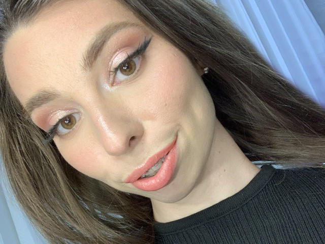 ChristieStorm has brown eyes, pink eyeshadow, black eyeliner, shiny pink lips, and a slight smile. She's wearing a black ribbed top, with long brown hair.