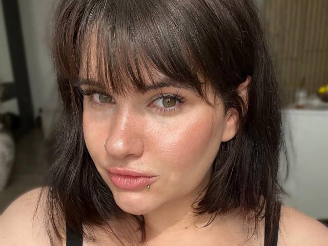 TinaColladas has dark brown, shoulder-length hair, green eyes, and a pierced lower lip. She's wearing a black top and has a nose ring. Her skin is slightly shiny.