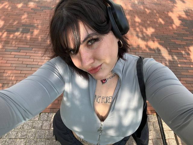 TinaColladas with short black hair, wearing headphones, a grey zip-up shirt, showing her tits, and a necklace, taking a selfie, looking up at the camera with a slight smile.