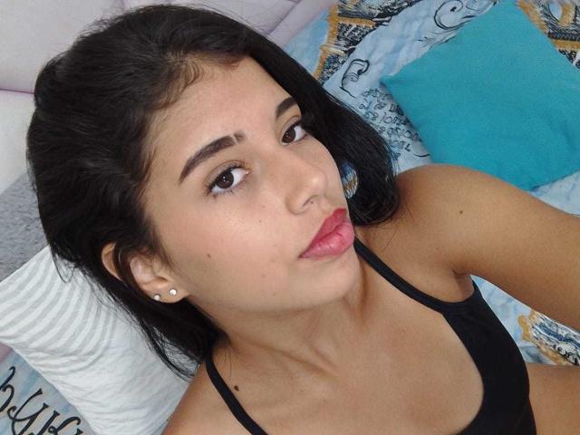 BarelyLegel has dark hair, brown eyes, and pink lipstick. She's wearing a black tank top, showing small tits, and has pearl earrings. She's looking up with a pouty face.