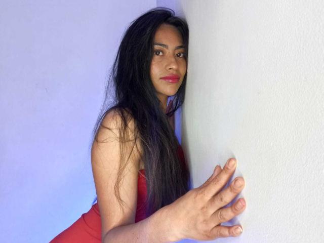 EmilyMitchell with long black hair, red dress, and pink lipstick, leaning against a white wall, right hand on the wall, looking at the camera with a slight smile.