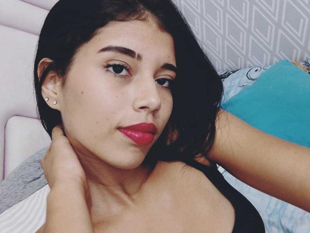 Ragazza con pelle media, capelli neri, rossetto rosso, canotta nera, orecchini, mano al collo, che guarda la fotocamera, con un sorriso leggero, carina, ambientazione: camera da letto..