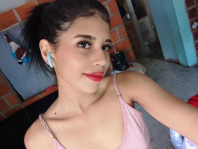 BarelyLegel has pale skin, dark hair in a ponytail, blue earbuds, pink top, red lipstick, small dark mole on her chest, and a slight smile, looking at the camera.