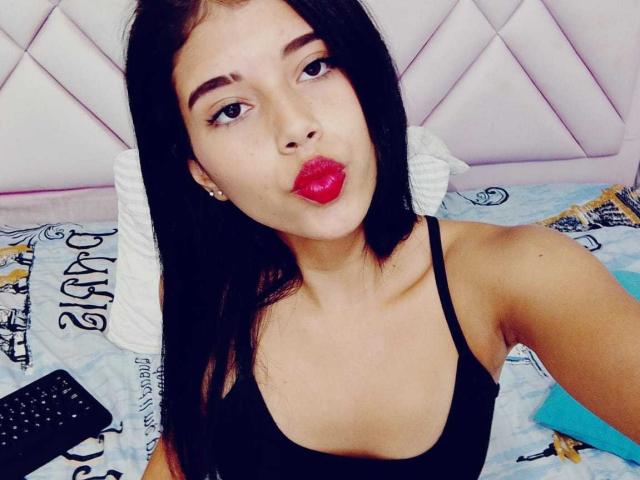 BarelyLegel, con i capelli neri, il rossetto rosso e gli occhi marroni, che fa la smorfia, indossa una canotta nera ed è seduta su un letto con un telecomando..