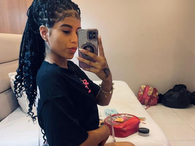 BarelyLegel, con lunghe trecce nere, pelle abbronzata e una maglietta nera, sta scattando un selfie allo specchio, mostrando un tatuaggio sulla mano e una trousse rossa sul letto..