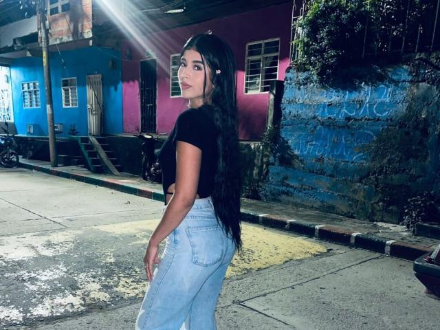 BarelyLegel, con lunghi capelli neri, una maglietta nera e jeans a vita alta color azzurro chiaro, è in piedi in strada di notte, guardando di spalle, mostrando il sedere..