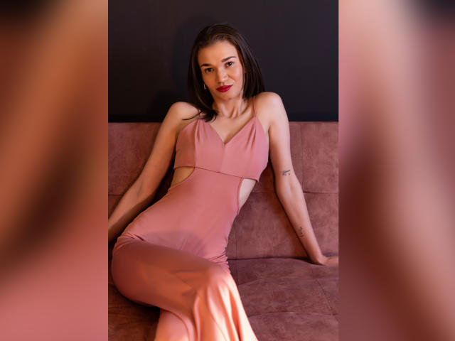 JennaDerose trägt ein pinkes Kleid mit Ausschnitten und sitzt auf einem pinken Sofa, die Beine gekreuzt, die Arme hinter ihr, mit rotem Lippenstift, Tätowierungen am rechten Arm, braunen Haaren und einem sanften Lächeln..