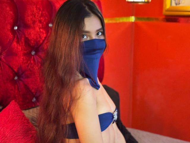 SweetlLove69 with long red-brown hair, blue mask covering mouth, blue strapless top, looking at camera, red tufted headboard, red wall, red pillow, black jacket, yellow frame.