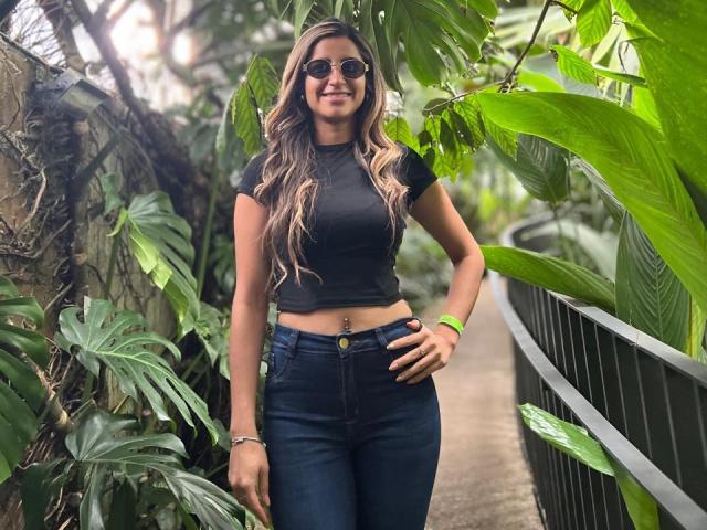 SofyaLane with long wavy brown hair, wearing a black crop top, blue jeans, and dark sunglasses, standing in a lush green tropical area, smiling with her hand on her hip.