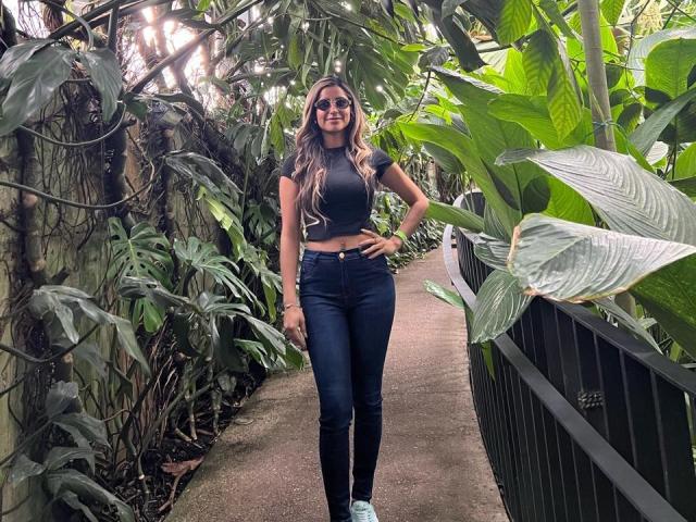 SofyaLane, avec de longs cheveux bruns, des lunettes de soleil, un crop top noir et un jean bleu moulant, se tient sur un chemin dans une serre, souriante, les mains sur les hanches, entourée de plantes vertes..