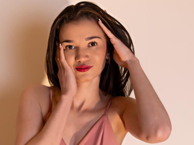 JennaDerose with hands on her face, pink dress, red lipstick, and brown hair against a plain background, showing her tits and a cute smile. Soft, warm lighting.