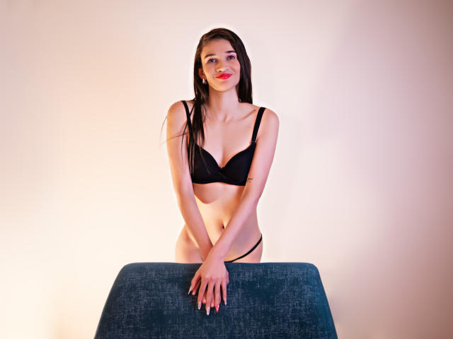 JennaDerose met lang zwart haar, rode lippenstift, een zwarte bh en een zwarte string, staat met haar handen op een blauwe stof, glimlachend, en laat kleine borsten en een slanke, sexy achterkant zien..