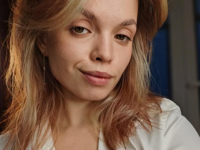 Sinteas has light brown eyes, subtle makeup, and a slight smile, with blonde and brown hair, wearing a white shirt, showing soft, natural tits and a cute, confident face.