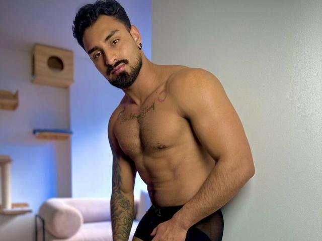 Muscular IvannaAndFriend with tan skin, dark curly hair, and beard, wearing black mesh pants, standing against a gray wall, showing tattoos and a hairy chest.
