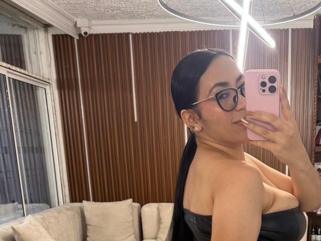 CamilleJuicyHot with black hair, glasses, and earrings, taking a mirror selfie in a leather top, showing her tits, with a pink phone, in a room with wood panels.