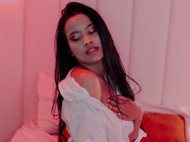 KarolPalacios with wet black hair, pink eye makeup, white shirt slipping off, hand on chest, red and orange lighting, slightly open mouth, looking down, sexy, soft pink background.