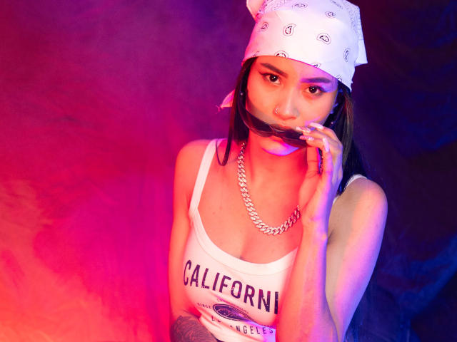 KarolPalacios, com uma bandana branca, piercing no nariz, colar de corrente, camiseta branca e iluminação rosada, segurando um cigarro perto da boca, mostrando tatuagens no braço..