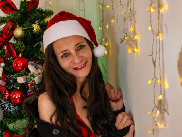 Meredith Hunnt, avec de longs cheveux bruns, du rouge à lèvres, souriante, portant un bonnet de Père Noël, un haut noir à épaules dénudées, une écharpe rayée rouge et noire, à côté d'un sapin de Noël et de guirlandes lumineuses jaunes..