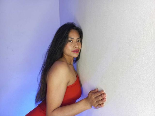 EmilyMitchell with long black hair, wearing a red strapless dress, standing against a white wall, hand on the wall, smiling slightly, showing her tits, with a blue light.