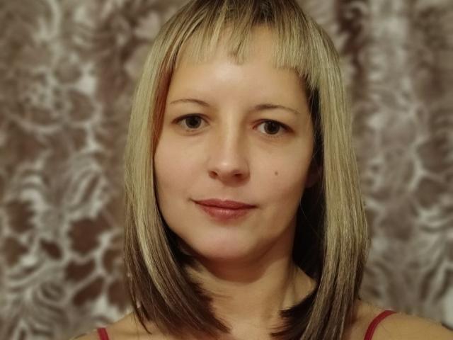 A portrait of AngelaRoxyx with straight, shoulder-length blonde hair featuring darker roots. She has light skin, brown eyes, and a small black mole on her right cheek. She wears a red sleeveless top. The background is a blurred, floral-patterned curtain. AngelaRoxyx's hair is smooth with a slight shine, and her expression is neutral. The lighting is even, highlighting her facial features. The overall composition is centered and close-up.