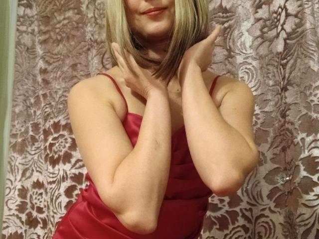 AngelaRoxyx with blonde hair, smiling, hands near neck, wearing a red satin dress, showing her arms and tits. She's cute and has a playful expression.