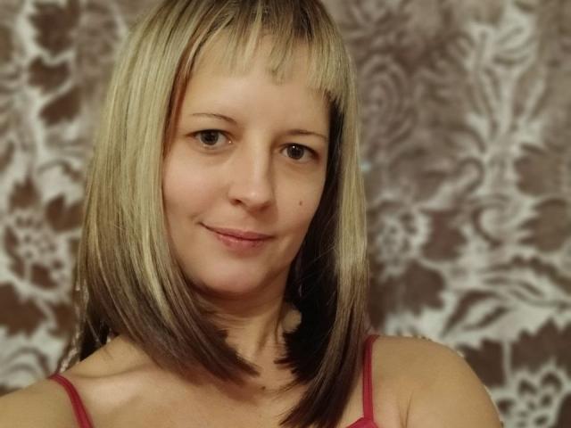 AngelaRoxyx with blonde and brown shoulder-length hair, small smile, brown eyes, red spaghetti strap top, showing a cute mole on her right cheek.