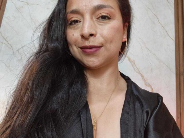 StangelarRivera with long black hair, tan skin, small beauty mark on left cheek, subtle smile, wearing black silk top, gold necklace, and gold earrings.